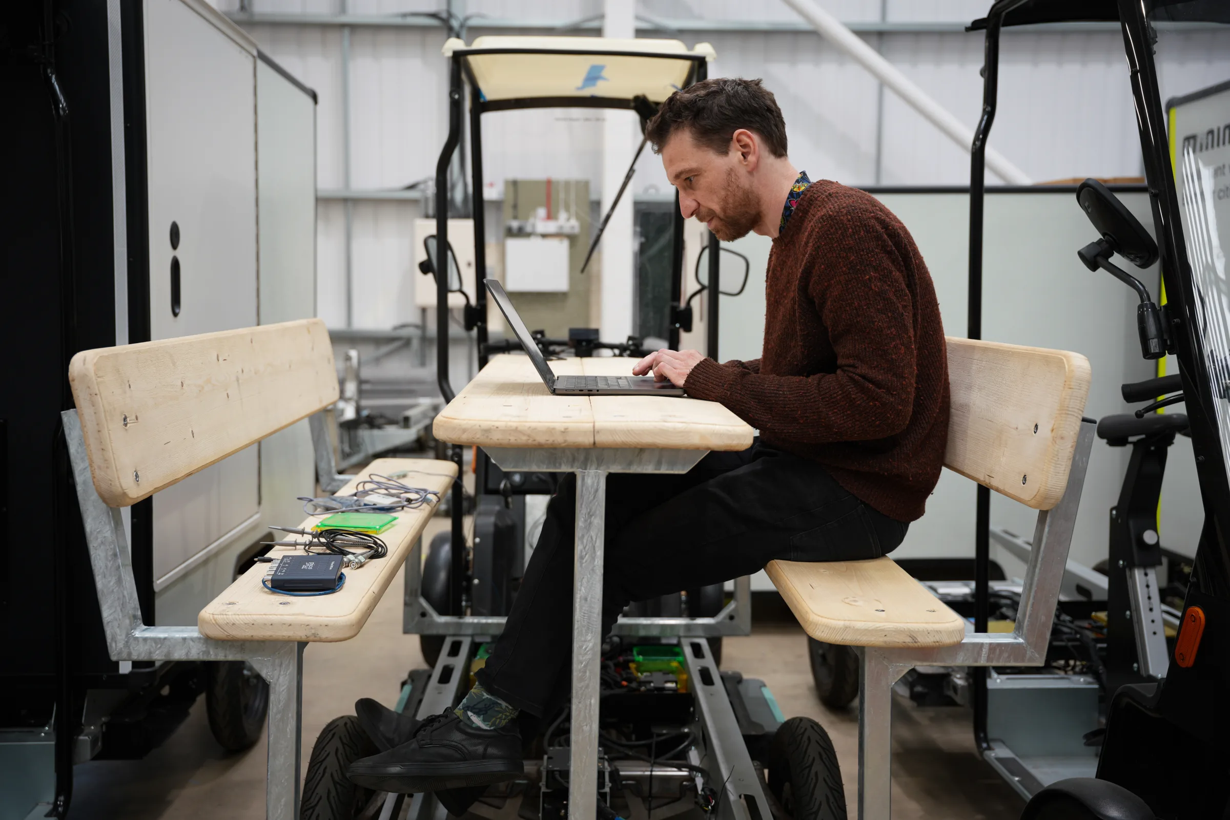 A Minimal engineer working on a laptop at a wooden bench built onto a vehicle chassis in the workshop