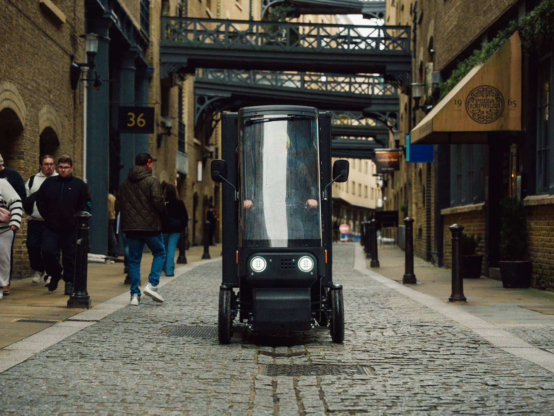 A Minimal Pedal 4 driving towards the camera along a cobbled London street, under an iron railway bridge, with pedestrians on the pavement beside it