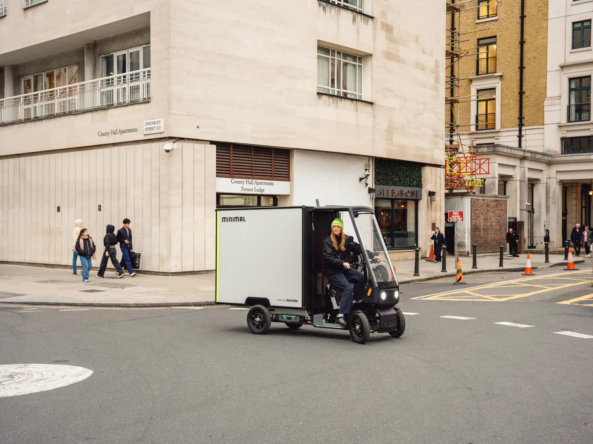 A Minimal box-body delivery vehicle turning a corner on a London street in front of a stone apartment building, with pedestrians walking past