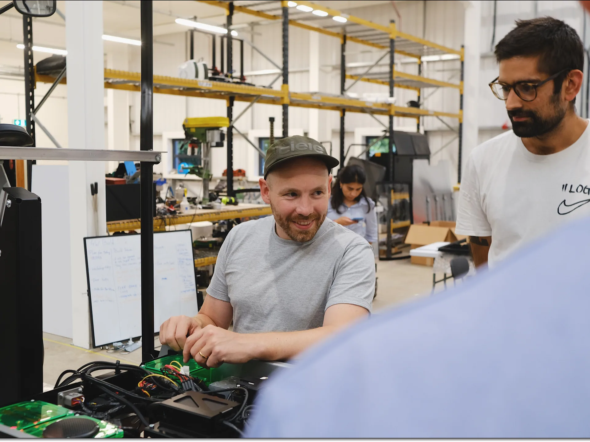 Two Minimal engineers at a workbench in the factory, one seated working on an exposed circuit board and the other standing beside him