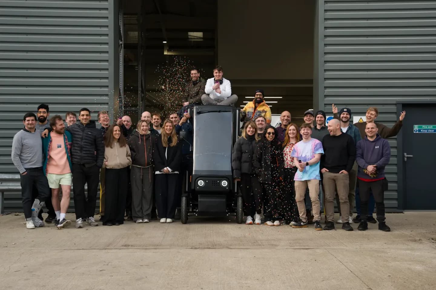 The Minimal team standing together in front of an open factory door, waving, with a Pedal 4 vehicle in the middle of the group